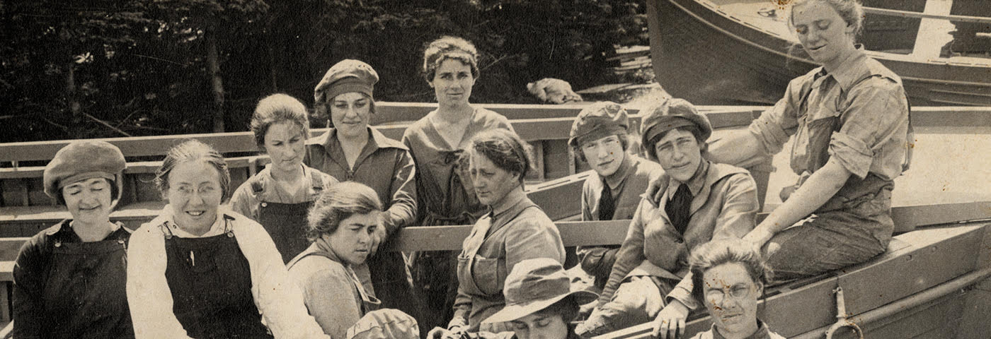 Women sitting in and standing in front of a lifeboat. There is another lifeboat behind them and a stand of trees to the left. The women are wearing work shirts, overalls, and matching wool hats.