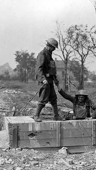 Image is a black and white photograph of two Canadian soldiers emerging from a German underground tunnel in France. One soldier is standing and lending his hand to the other soldier, who is exiting the tunnel. There are bombed out buildings in the background.