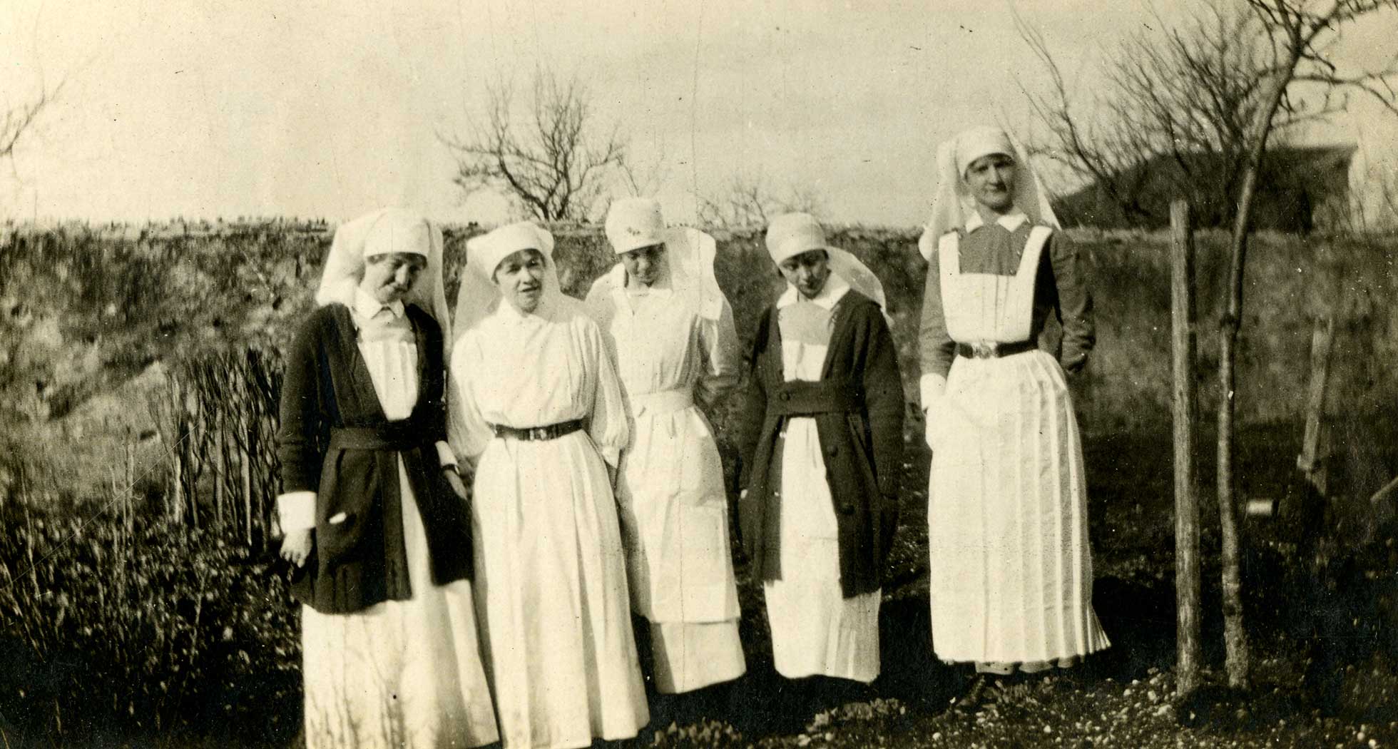 Photographie en noir et blanc de cinq infirmières militaires en uniforme, debout à l’extérieur devant un mur.