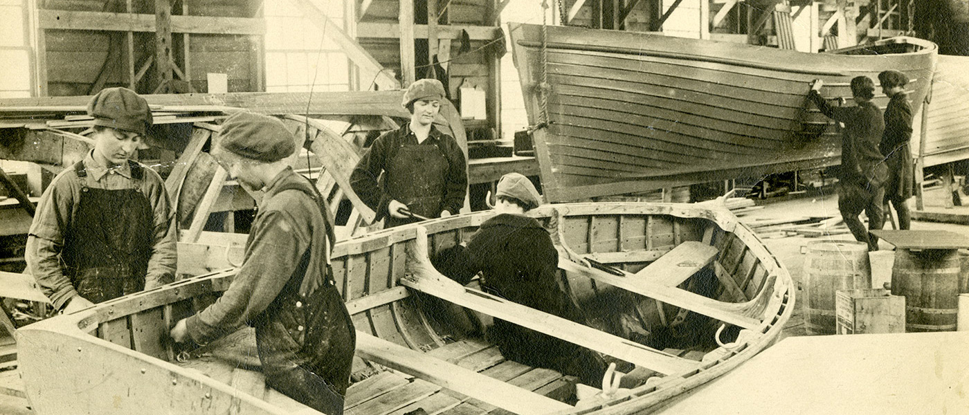 Sepia photograph of four women working on a lifeboat in a large warehouse. Two women are in the background of the photograph inspecting a boat hung from the ceiling. The women are wearing work shirts, overalls, and matching wool hats.
