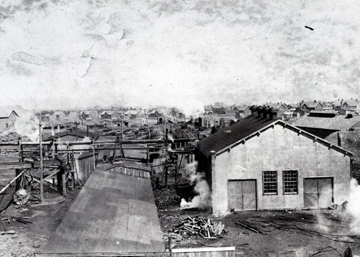 Photographie en noir et blanc d’un paysage industriel dans une ville de charbonnage, avec au loin des maisons fournies par la compagnie. On peut voir à gauche des trains qui transportent du charbon.