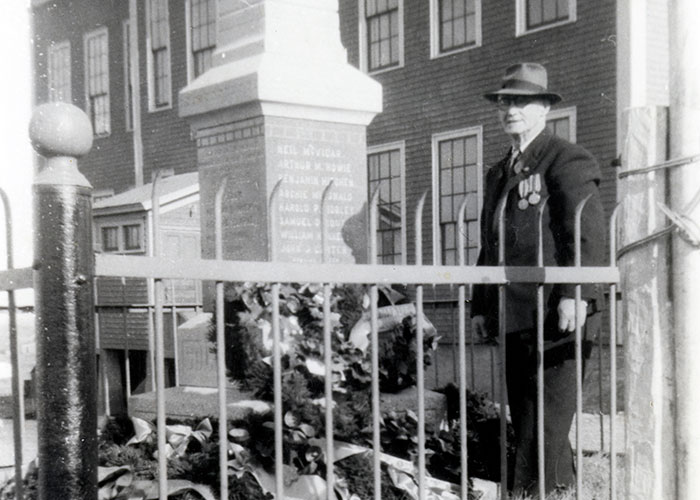 Photographie en noir et blanc d’un homme debout près d’un cénotaphe érigé en mémoire de la guerre. Il y a des couronnes autour du cénotaphe et une clôture entoure le périmètre du cénotaphe.