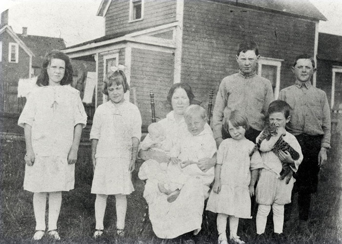Black and white photograph of eight people standing outside coal company houses. The mother is sitting centre, with a young boy on her lap. The two older daughters are standing left of her and the old eldest boys at the right in the back. Two young children are to her right, with a boy holding a pet cat.