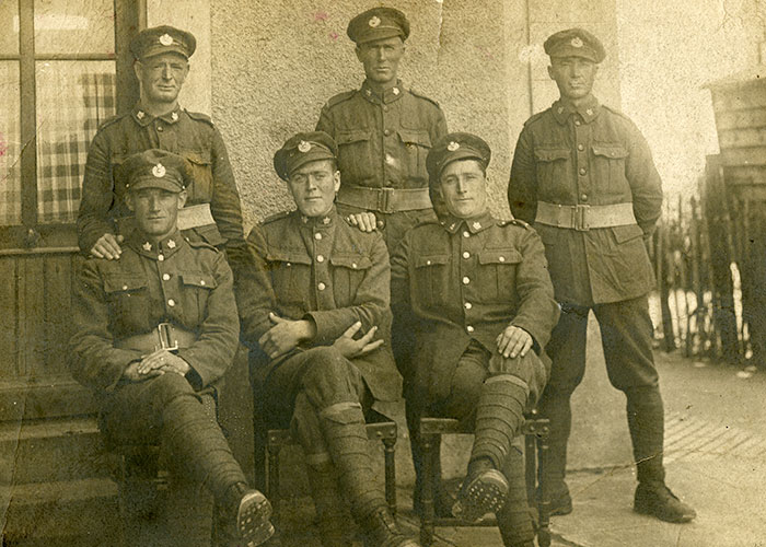 Sepia photograph of six military men in uniform. Three men are sitting on a bench outdoors with their legs crossed. Three men are standing behind them.